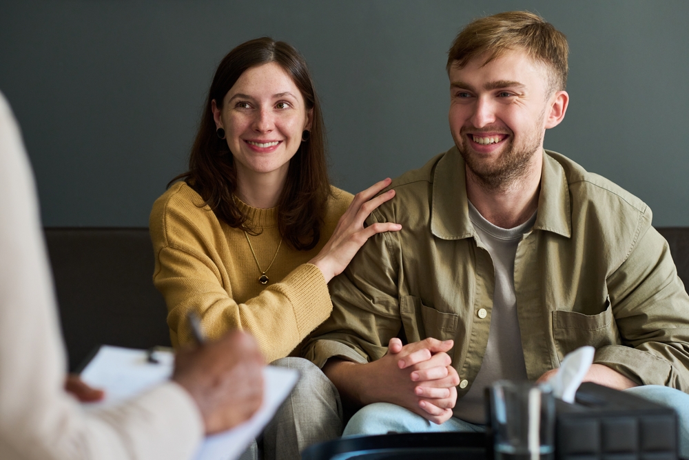 Caucasian young adult woman and Caucasian young adult man sitting together holding hands smiling while attending counseling session with professional writing on clipboard